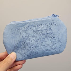 May include: A light blue zippered pouch featuring a dark blue line art illustration of a bakery interior. The design includes shelves filled with various baked goods, a counter, and a banner at the top with the text FRESH BREAD.