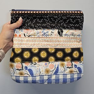 May include: A quilted clutch with a navy blue zipper and a variety of patterned fabric panels. The panels include black fabric with white birds, floral prints, and a black panel with yellow suns. The bottom panel is blue and white checkered.