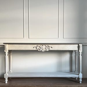 May include: A cream-coloured console table with ornate carvings and a lower shelf. The table features decorative moulding and is supported by four fluted legs. The tabletop is a contrasting grey colour. The table is approximately 152 cm long.
