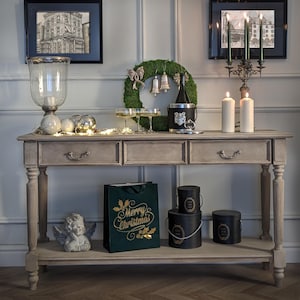 May include: A decorated console table with Christmas decorations. A green wreath with bells, champagne glasses, and a bottle of champagne in an ice bucket are on the table. A gift bag with "Merry Christmas" text and black hat boxes are also present. The table is grey.