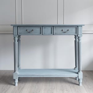 Può includere: Una consolle azzurra con due cassetti e un ripiano inferiore. Il tavolo ha gambe scanalate ornate e maniglie dei cassetti in metallo curve. Il design è classico ed elegante, adatto per un ingresso o un soggiorno.