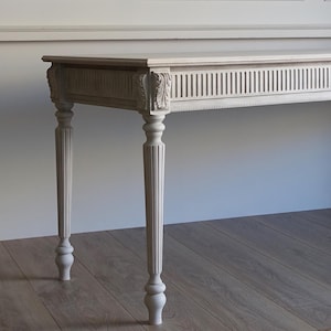 May include: A light gray console table featuring a rectangular top and four tapered legs with vertical fluting. The apron is decorated with a repeating vertical groove pattern and carved leaf motifs at the top of each leg, finished in a matte tone.