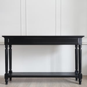May include: A rectangular black console table featuring a smooth top surface, a lower shelf, and four fluted legs with decorative carved detailing. The furniture piece has a solid matte finish and a structured frame with clean, straight lines.