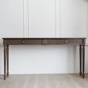 Può includere: Una lunga consolle in legno marrone scuro con due cassetti e ferramenta circolare decorativa. Il tavolo ha gambe sottili e scanalate e un design intagliato dettagliato sul davanti. Lo stile generale è classico ed elegante.