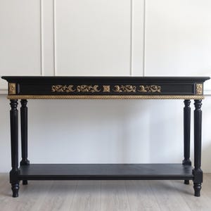 Puede incluir: Una mesa consola negra con tallas ornamentales de color dorado a lo largo del frente y las esquinas. La mesa tiene cuatro patas estriadas y un estante inferior. La parte superior rectangular está acabada en negro, con un borde dorado trenzado.