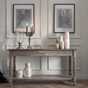 Peut inclure: Une console grise avec un plateau en bois, ornée d'objets décoratifs. Deux croquis en noir et blanc encadrés sont accrochés au-dessus. La table présente une lanterne en verre, un chandelier avec des bougies vertes et des vases décoratifs.