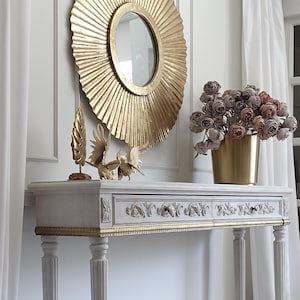 May include: A gold sunburst mirror hangs above a grey console table with gold trim. The table displays a gold bird sculpture and a gold vase filled with pink and brown flowers. White curtains frame the scene.