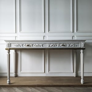 May include: A light gray console table featuring two drawers with intricate floral carvings, a dark metallic trim along the apron, and four fluted, tapered legs. The rectangular piece has a smooth top surface and small round drawer knobs.