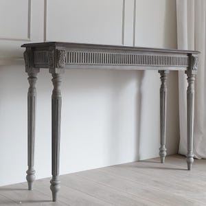May include: A gray console table featuring a rectangular top, a long apron with vertical fluted detailing, and carved leaf motifs at the corners. The table is supported by four slender, turned legs with vertical grooves and rounded feet.