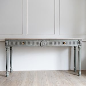 May include: A gray-blue console table with a light wood top. The table features two drawers with brass knobs and a central decorative circular element. The legs are fluted and painted in the same gray-blue color. The table is set against a white wall.