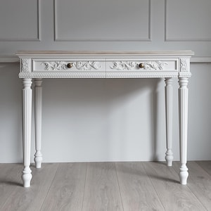 May include: A white console table featuring a light wood top, two drawers with intricate floral carvings, and two small round brass knobs. The table stands on four fluted legs with decorative square blocks at the top corners and a beaded trim detail.