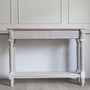 Peut inclure: Une console grise claire avec un dessus en bois naturel. La table présente des détails sculptés sur les pieds et le tablier, avec une étagère inférieure. La table mesure environ 122 cm de long.