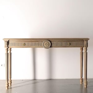 Könnte beinhalten: Ein hellbrauner Holzkonsolentisch mit dekorativem Schnitzdesign. Der Tisch hat ein zentrales kreisförmiges Medaillon, vertikale Lamellen und verzierte Details an den Beinen und Ecken. Der Tisch ist etwa 152 cm lang.