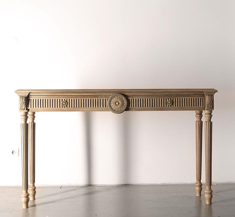 Op de afbeelding: Een lichtbruine houten console met een decoratief gesneden ontwerp. De tafel heeft een centraal cirkelvormig medaillon, verticale lamellen en sierlijke details op de poten en hoeken. De tafel is ongeveer 152 cm lang.