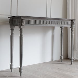 以下が含まれることがあります： 長方形の天板と4本の回転脚を備えたグレーの木製コンソールテーブル。テーブルは前面にスラットデザインがあり、コーナーには彫刻が施されています。テーブルは落ち着いたグレーの色で塗装されています。