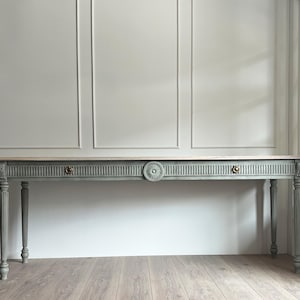 May include: A long, rectangular console table with a muted sage green finish. The table features a light wood top, fluted legs, and a decorative central rosette medallion. Two small, round brass-toned drawer pulls are positioned on the front.