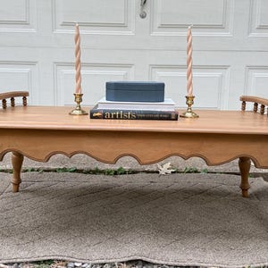 May include: A light brown wooden coffee table with curved legs and a scalloped edge. Two twisted candles in gold candle holders, a blue box, and books are on the table. The book title is "artists".