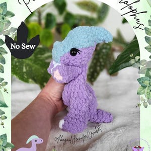 May include: A handmade, purple crochet dinosaur toy with a light blue crest. The toy has black eyes and pink accents. The text "Penelope The Parasaurolophus" is above the dinosaur. The words "No Sew" are also visible.