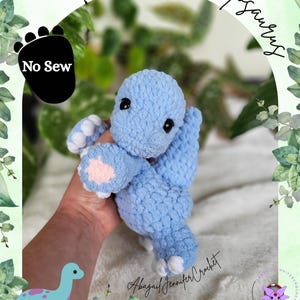 Peut inclure: Une peluche Brontosaurus bleu clair crochetée avec des détails blancs et roses. Le jouet a des yeux noirs et est tenu dans une main. L'image comprend le texte "Blain The Brontosaurus" et "No Sew".
