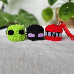 May include: Three crochet keychains: a green figure with black eyes, a black figure with purple eyes, and a red TNT block with gray and white accents. Each keychain has a silver ring and a red tassel.