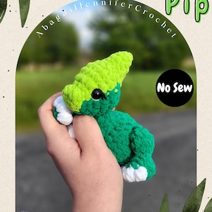 Puede incluir: Un juguete de dinosaurio de ganchillo verde y blanco hecho a mano llamado Pip. El dinosaurio tiene una cabeza verde lima y un cuerpo verde oscuro. El texto "Tiny Tots Pip" y "No Sew" son visibles.