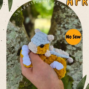 May include: A small, plush, crocheted dinosaur toy featuring a mustard yellow body, a light blue head and back plate, and white feet. The text Tiny Tots Ark, AbagailJenniferCrochet, and No Sew is visible on the graphic overlay surrounding the toy.
