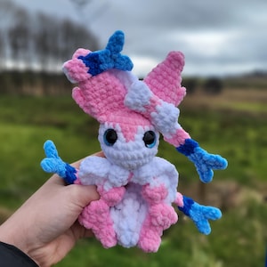 Op de afbeelding: Een handgemaakte gehaakte Sylveon-knuffel in roze, wit en blauw. De knuffel heeft grote blauwe ogen en gedetailleerde kenmerken, waaronder linten en antennes. De knuffel wordt vastgehouden voor een vage buitenachtergrond.