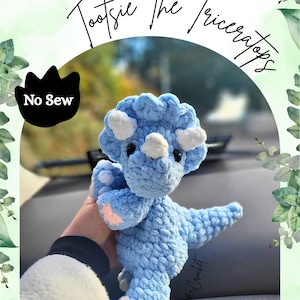 Può includere: Un giocattolo a forma di dinosauro triceratopo all'uncinetto blu chiaro con dettagli bianchi. Il giocattolo ha un aspetto strutturato ed è etichettato "Tootsie The Triceratops" e "No Sew". Il giocattolo è tenuto davanti a uno sfondo sfocato.
