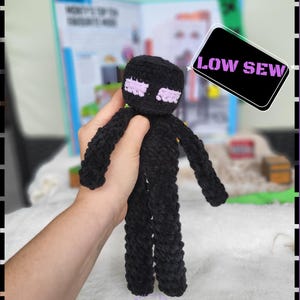 May include: A black crochet Enderman doll with lavender eyes and outstretched arms. The doll is held in a person's hand. The image includes the text "ENDERBOI" and a "LOW SEW" label.