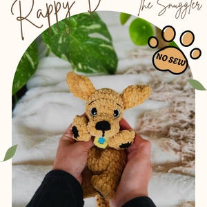 Op de afbeelding: Een gehaakte bruine hondenknuffel met zwarte poten en een blauwe kraag wordt in handen gehouden. De knuffel heeft grote oren en een kleine staart. De tekst "Rappy Doo The Snuggler" staat boven de hond. De afbeelding bevat ook een "No Sew"-afbeelding.