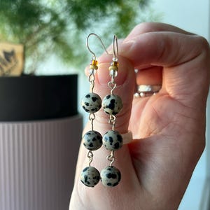 May include: Dangle earrings with three cream-coloured, speckled, round beads with black spots. The earrings have silver-coloured hooks and small gold and clear beads. The earrings are held up by a hand.