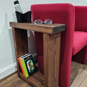 May include: A dark brown wooden side table with a rectangular top and a lower shelf. A black remote holder and glasses rest on top. Two books are on the shelf. A red chair is in the background.