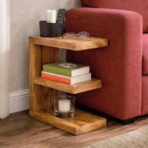 May include: A three-tiered wooden side table with a natural finish. The table holds a black container with a candle and remote, glasses, two books, and a candle in a glass. The table is next to a red sofa.