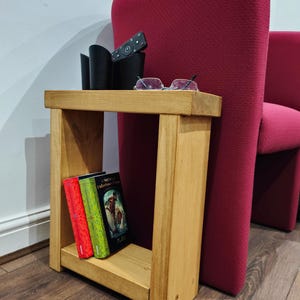May include: A small, light-coloured wooden side table with a lower shelf holding three books. A black remote and glasses rest on the tabletop. A red armchair is in the background.