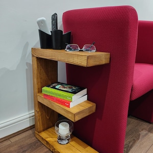 May include: A three-tiered wooden side table with a warm, honey-toned finish. The top shelf holds a black organizer with remotes and glasses. The middle shelf has two books, and the bottom shelf has a candle in a glass holder.