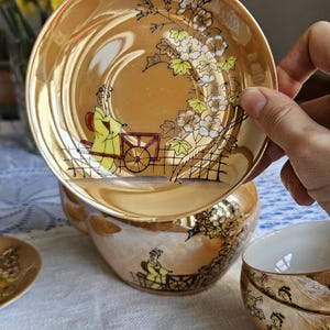 May include: Vintage tea set with a gold luster finish. The set includes a teacup, saucer, and a small cup. Each piece is decorated with a hand-painted scene of a person in a yellow robe with a cart, and floral accents.