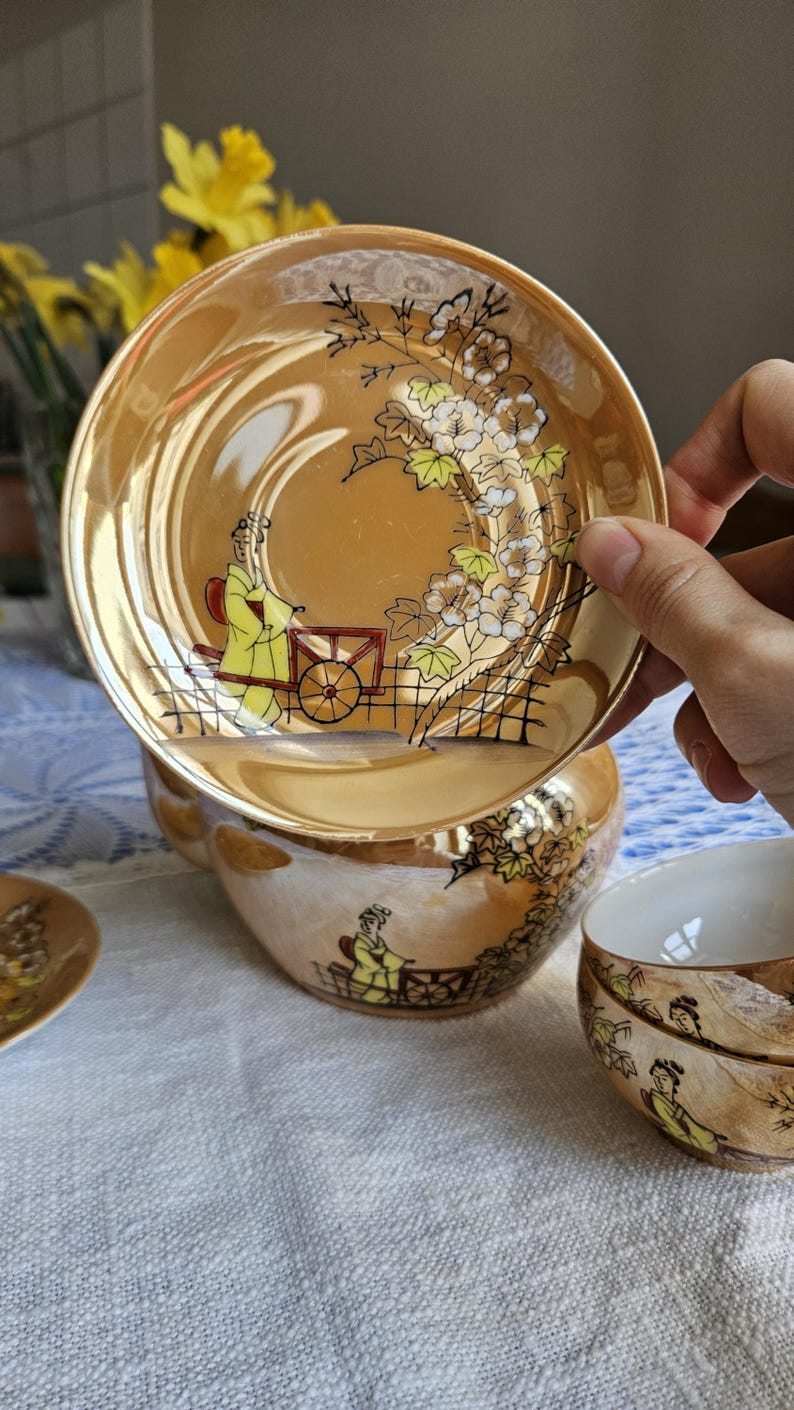 May include: Vintage tea set with a gold luster finish. The set includes a teacup, saucer, and a small cup. Each piece is decorated with a hand-painted scene of a person in a yellow robe with a cart, and floral accents.