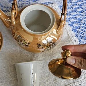 May include: A vintage tea set featuring a gold-toned teapot with a woven handle and floral accents. The set includes a white tea strainer and a lid with a gold knob. The teapot is on a white surface.