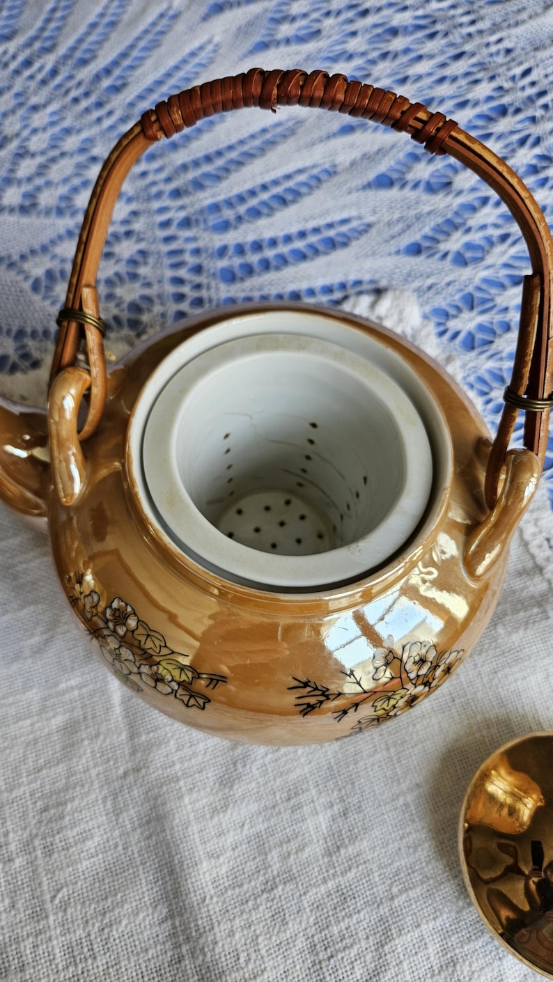 May include: A vintage, iridescent gold-colored teapot with a woven handle. The teapot features a floral design and a removable infuser. The teapot is sitting on a white cloth.