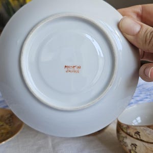 May include: A white saucer with a circular indentation, featuring the text "MADE IN JAPAN" in red. The saucer is part of a tea set, with a matching teacup visible. The background includes yellow flowers.