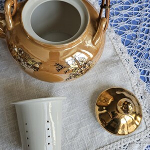 May include: A vintage tea set with a gold-toned teapot, a white tea strainer, and a gold lid. The teapot has a woven handle and floral designs. The set is on a white cloth with a lace trim, set against a blue background.