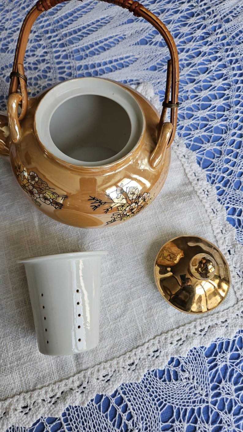 May include: A vintage tea set with a gold-toned teapot, a white tea strainer, and a gold lid. The teapot has a woven handle and floral designs. The set is on a white cloth with a lace trim, set against a blue background.