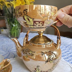 May include: A vintage tea set with a gold luster finish. The set includes a teapot, a teacup, and a saucer, all adorned with floral and oriental figures. The teacup is balanced on top of the teapot. The set is on a white tablecloth.