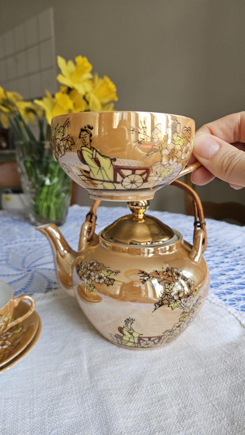 May include: A vintage tea set with a gold luster finish. The set includes a teapot, a teacup, and a saucer, all adorned with floral and oriental figures. The teacup is balanced on top of the teapot. The set is on a white tablecloth.