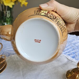 May include: A vintage, gold-toned teapot with a detailed illustration. The bottom of the teapot is white with the text "MADE IN JAPAN" in red. The teapot is part of a tea set, with a cup and saucer visible.