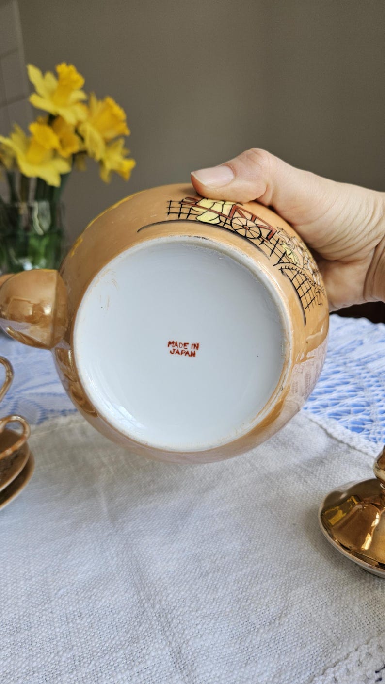 May include: A vintage, gold-toned teapot with a detailed illustration. The bottom of the teapot is white with the text "MADE IN JAPAN" in red. The teapot is part of a tea set, with a cup and saucer visible.
