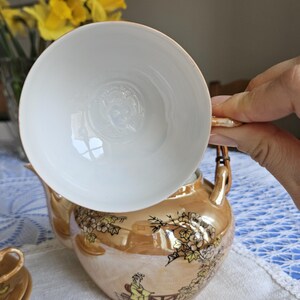 May include: A vintage tea set with a gold-toned teapot and a white teacup. The teapot features a painted design of a person and floral elements. The teacup is held above the teapot, showcasing its interior design. The set is on a white tablecloth.