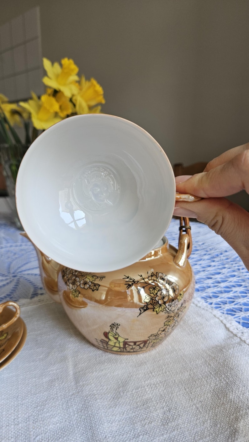May include: A vintage tea set with a gold-toned teapot and a white teacup. The teapot features a painted design of a person and floral elements. The teacup is held above the teapot, showcasing its interior design. The set is on a white tablecloth.