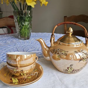 May include: A vintage tea set with a gold luster finish, featuring a teapot, two teacups, and saucers. The set is decorated with a floral and landscape design. A vase of yellow daffodils sits in the background.
