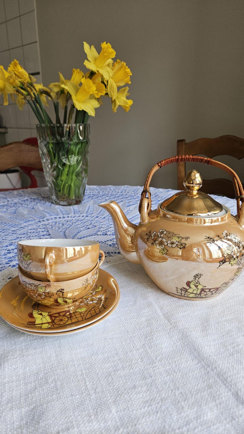 May include: A vintage tea set with a gold luster finish, featuring a teapot, two teacups, and saucers. The set is decorated with a floral and landscape design. A vase of yellow daffodils sits in the background.